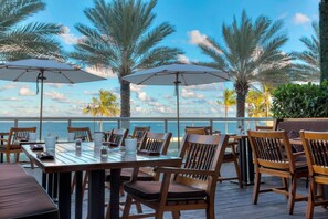 An outdoor dining area with wooden tables and chairs, umbrellas, and a view of the ocean. - The W - Bayshore Unit 1003 (Fort Lauderdale)
