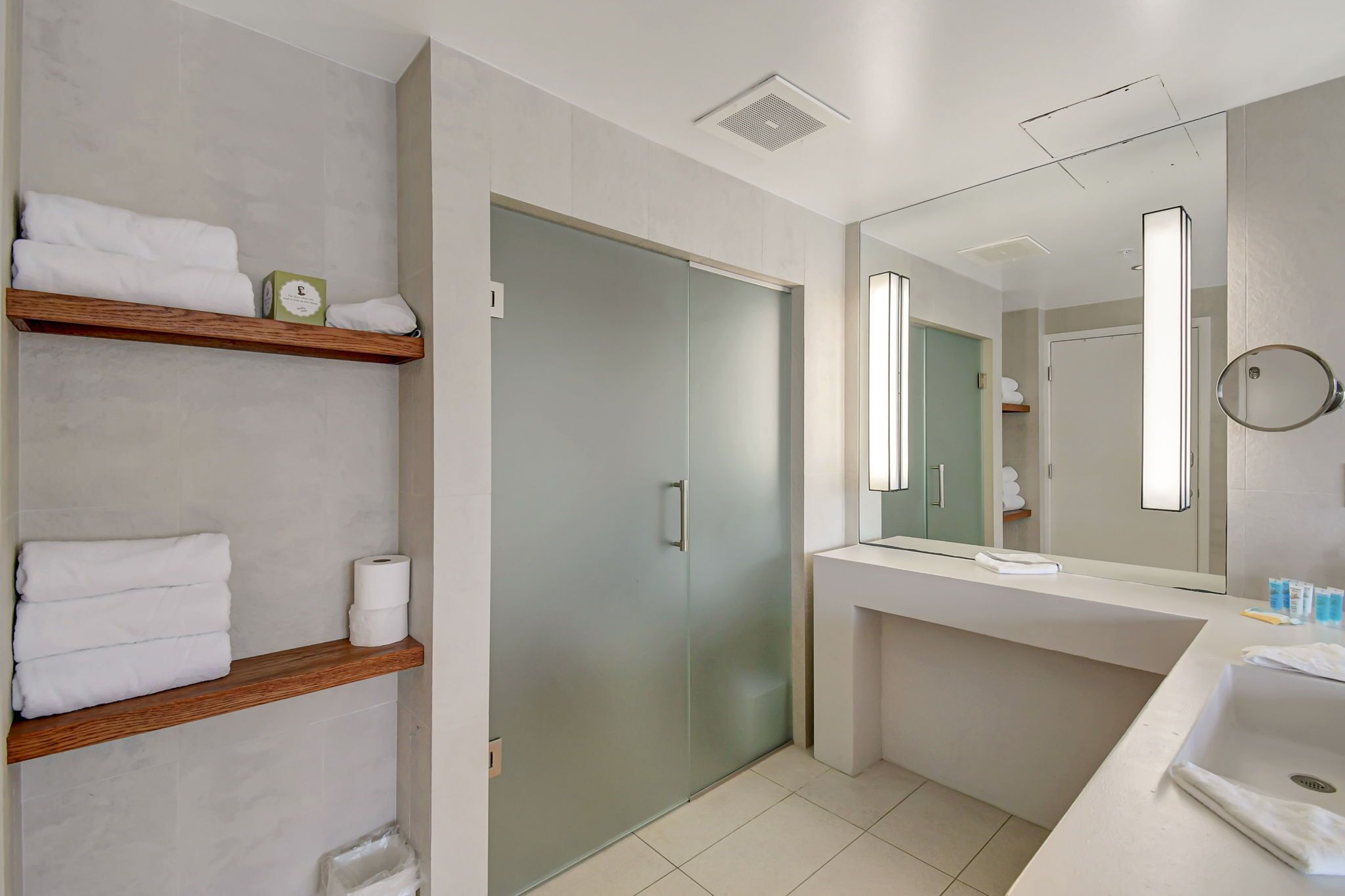 A modern bathroom with a glass shower door, a wooden shelf with towels, a large mirror, and a white sink.