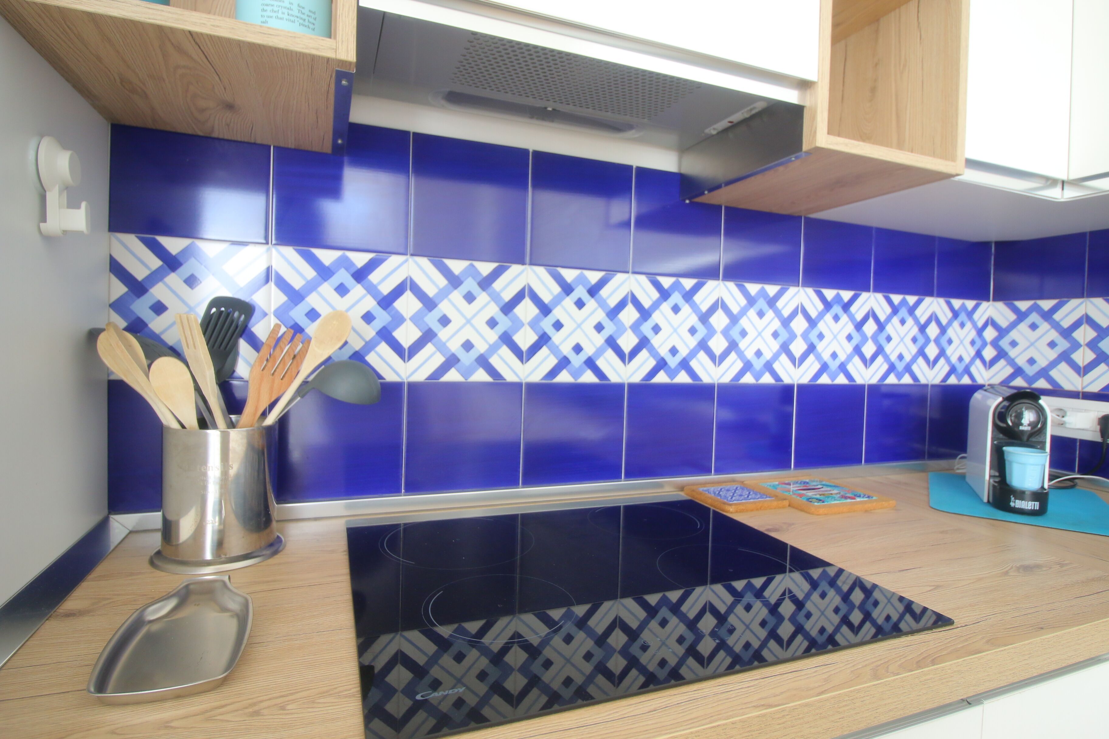 Una cocina con baldosas azules y blancas estampadas, un recipiente de acero inoxidable para utensilios, una placa de cocina por inducción negra y una cafetera.