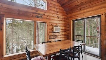 A log cabin with a wooden dining table and chairs, large windows, and a view of the forest.