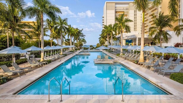 Zona de piscina con sillones de descanso y sombrillas, rodeada de palmeras y un edificio con varios balcones.