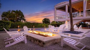 Un área de estar al aire libre moderna con sillas Adirondack blancas y una chimenea, rodeada por una piscina y palmeras.