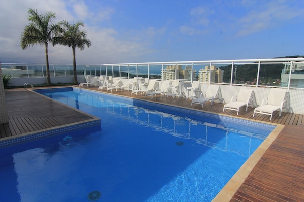 A rooftop pool with clear blue water, surrounded by white lounge chairs and palm trees.