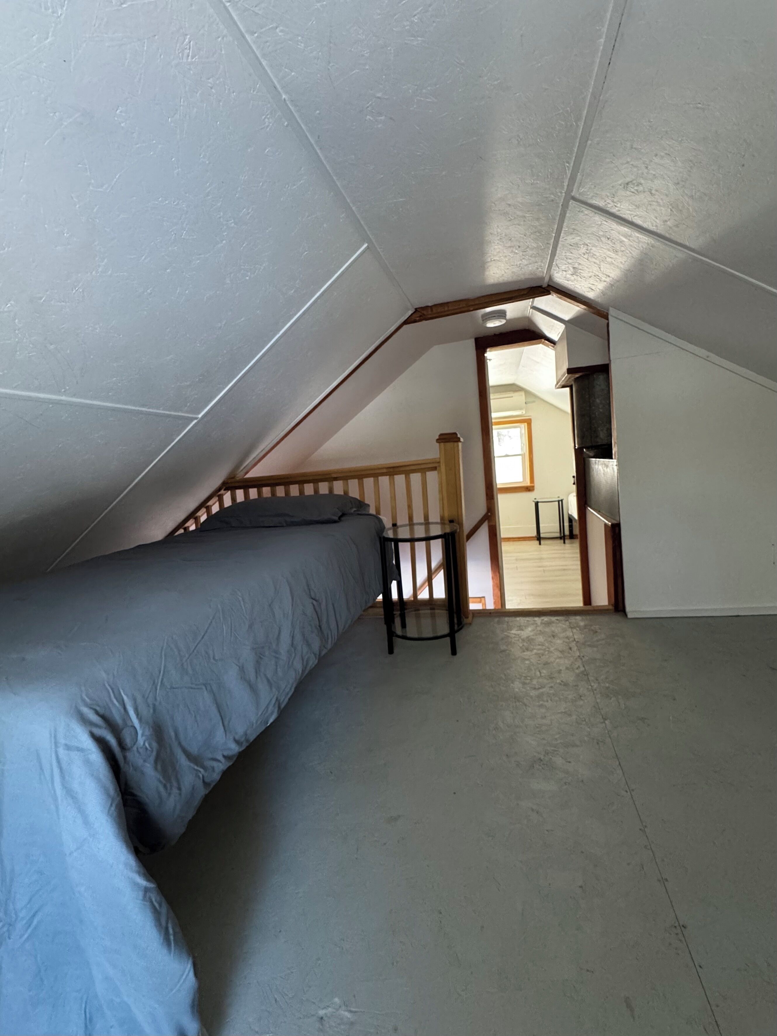 Quarto sótão pequeno com telhado inclinado, uma cama de solteiro, uma mesa e vista para a cozinha.
