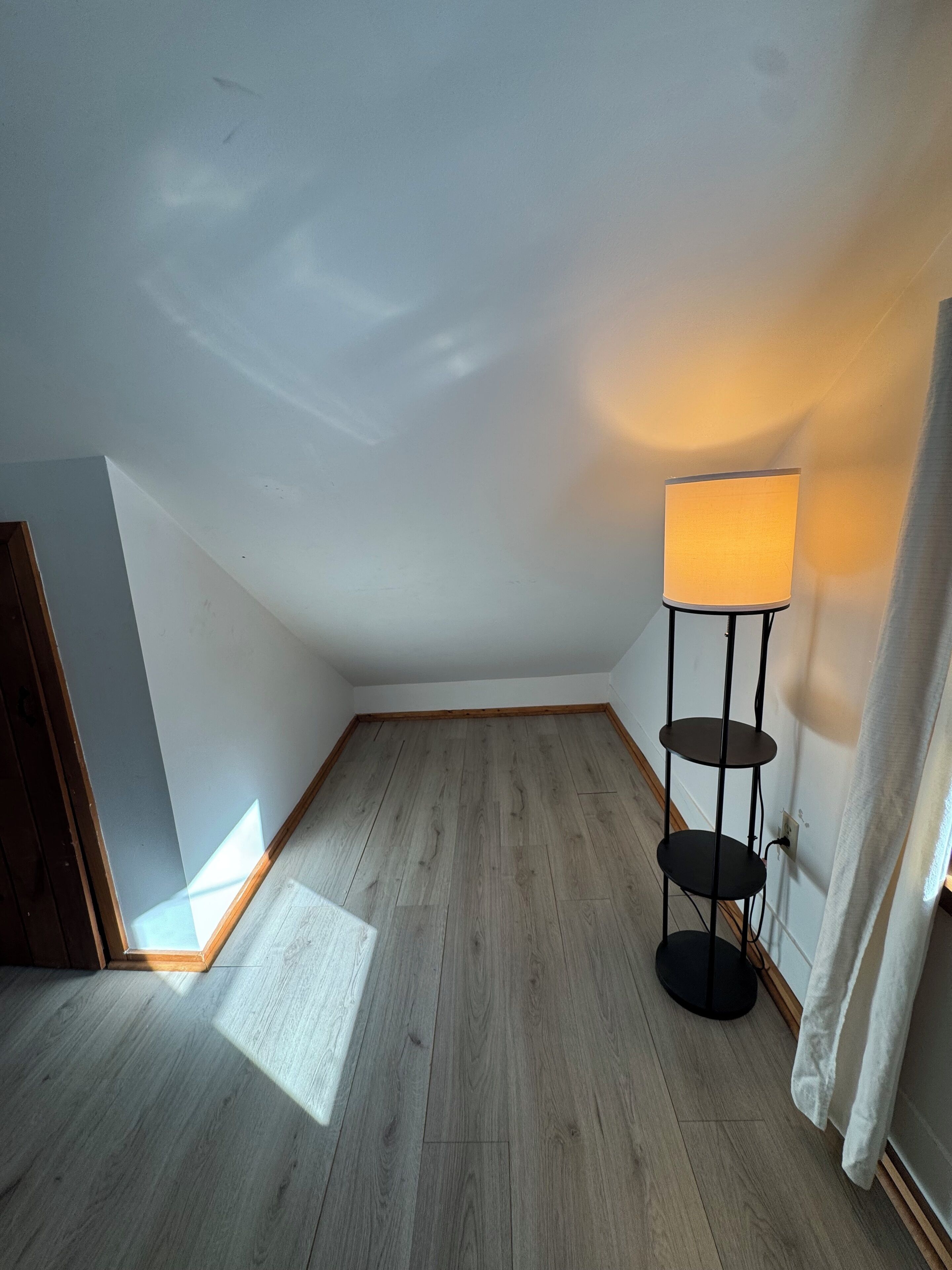 Una habitación con techo inclinado, suelo de madera, una lámpara de pie y una cortina.