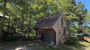 Un piccolo chalet in legno con tetto spiovente, un garage e circondato da alberi fitti.