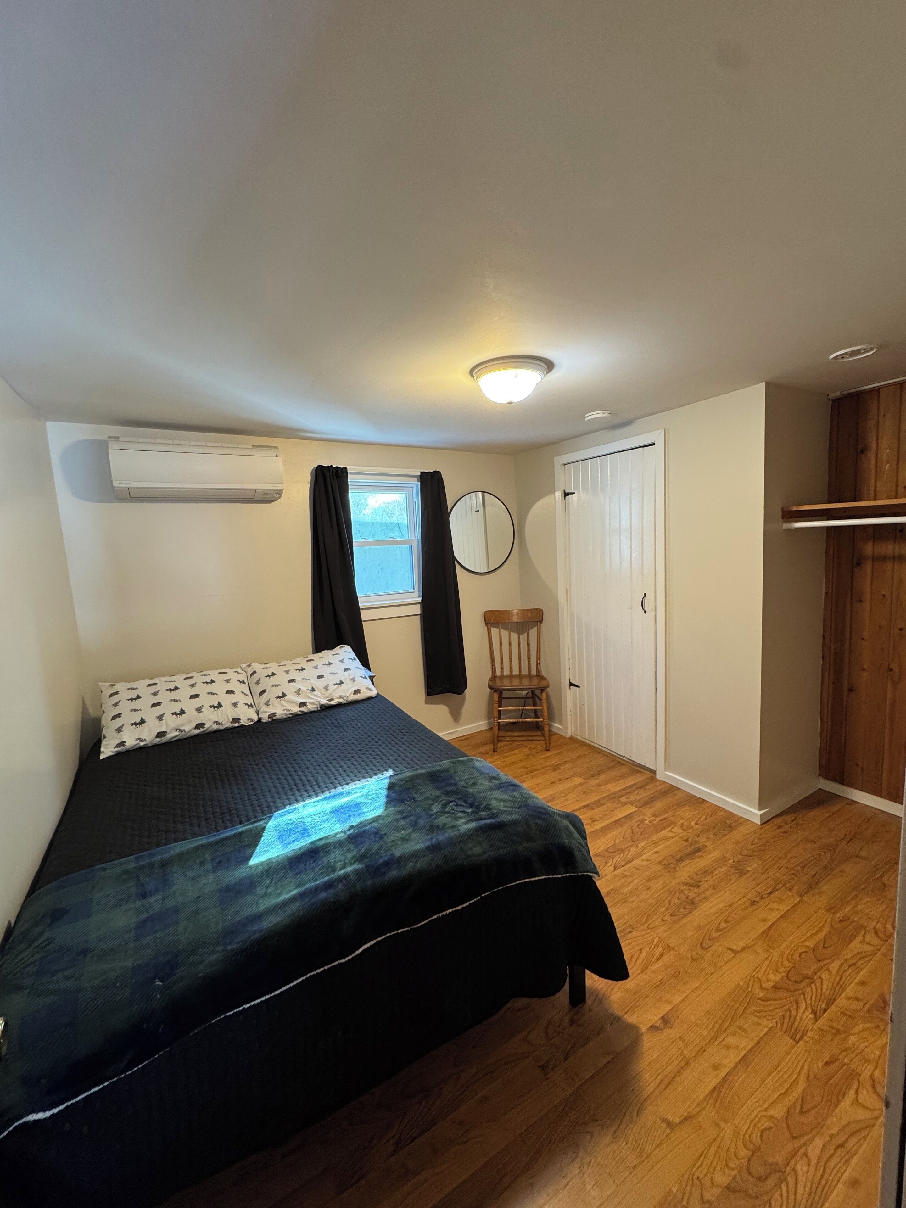 A bedroom with a bed, a chair, a wardrobe, a window, and an air conditioning unit.