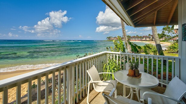 Een balkon met een witte reling, een ronde tafel en twee witte stoelen met uitzicht op een strand en de oceaan.