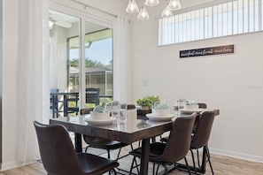 A dining area with a glass-top table, six chairs, and a sign on the wall. - Disney World, Universal Epic Waterfront Modern Home - Private Pool And Game Room (Kissimmee)
