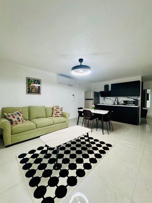 Sala de estar con sofá, una mesa de comedor y sillas. Hay una cocina con armarios y un refrigerador.