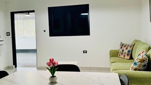 Ein modernes Wohnzimmer mit einem an der Wand montierten Flachbildfernseher, einem grünen Sofa mit gemusterten Kissen, einem Esstisch mit Stühlen und einer Vase mit rosa Blumen.