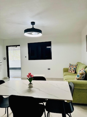 Una sala de estar moderna con un televisor de pantalla plana montado en la pared, un sofá verde con cojines estampados, una mesa de comedor con sillas y un jarrón con flores rosas.