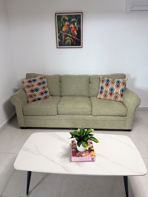 A green sofa with patterned pillows, a white table with a plant, and a framed painting above.