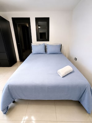 Habitación individual con una colcha azul claro, dos almohadas, una toalla blanca y un armario negro.