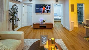 A modern living room with a flat-screen TV mounted on the wall, a wooden entertainment unit, a sofa, a coffee table with decorative items, and a glass shower enclosure visible through an open door. - Casa Kiwi in Miami - Sleeps 6 (Miami)