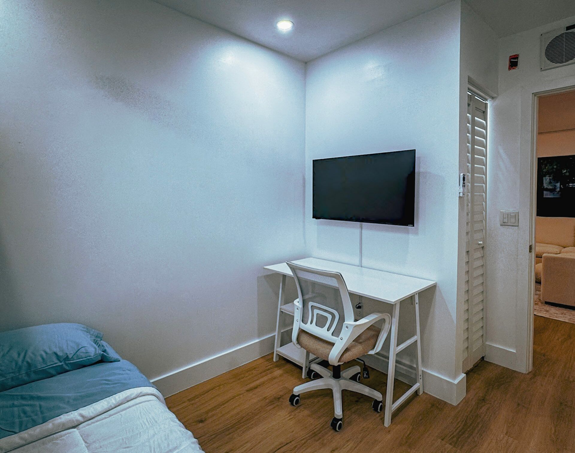 Une chambre comprenant un lit, un bureau avec une chaise, un téléviseur fixé au mur et une porte donnant sur une autre pièce.