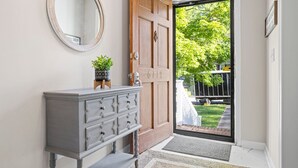 Un corridoio con una porta in legno, un comò grigio, uno specchio rotondo e vista su un giardino verde attraverso la porta.