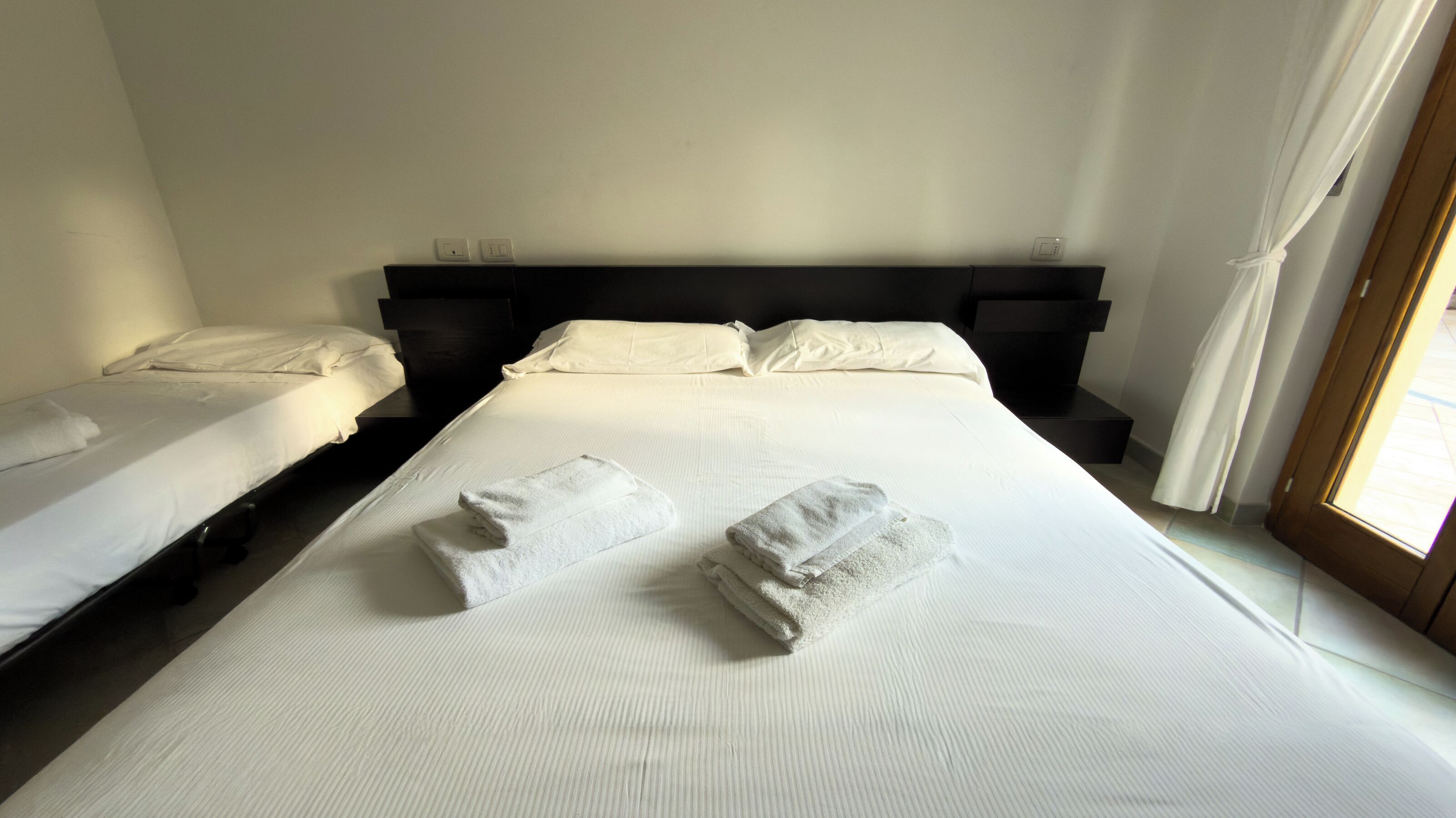 Een hotelkamer met twee eenpersoonsbedden, een groot bed met handdoeken, een zwart hoofdeinde en een raam met gordijnen.