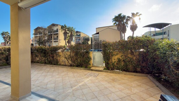 Un quartiere residenziale con patio piastrellato, ingresso chiuso da cancello e palazzi con balconi e palme.