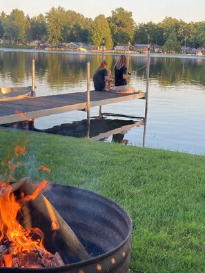 A lakeside dock with a fire pit in the foreground and two people sitting on the dock.