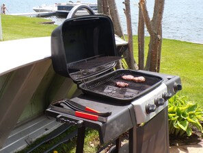 A portable grill with two burgers cooking, set on a grassy area near a body of water.