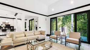 A modern living room with a beige sofa, a glass coffee table, and a view of greenery through large windows.
