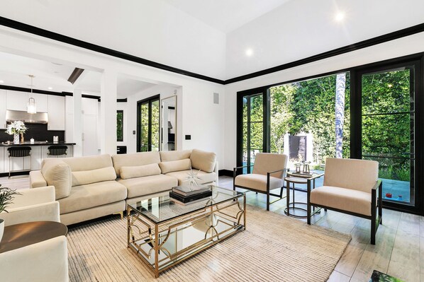 Un salón moderno con un sofá color beige, una mesa de centro de vidrio y vistas a zonas verdes a través de amplios ventanales.
