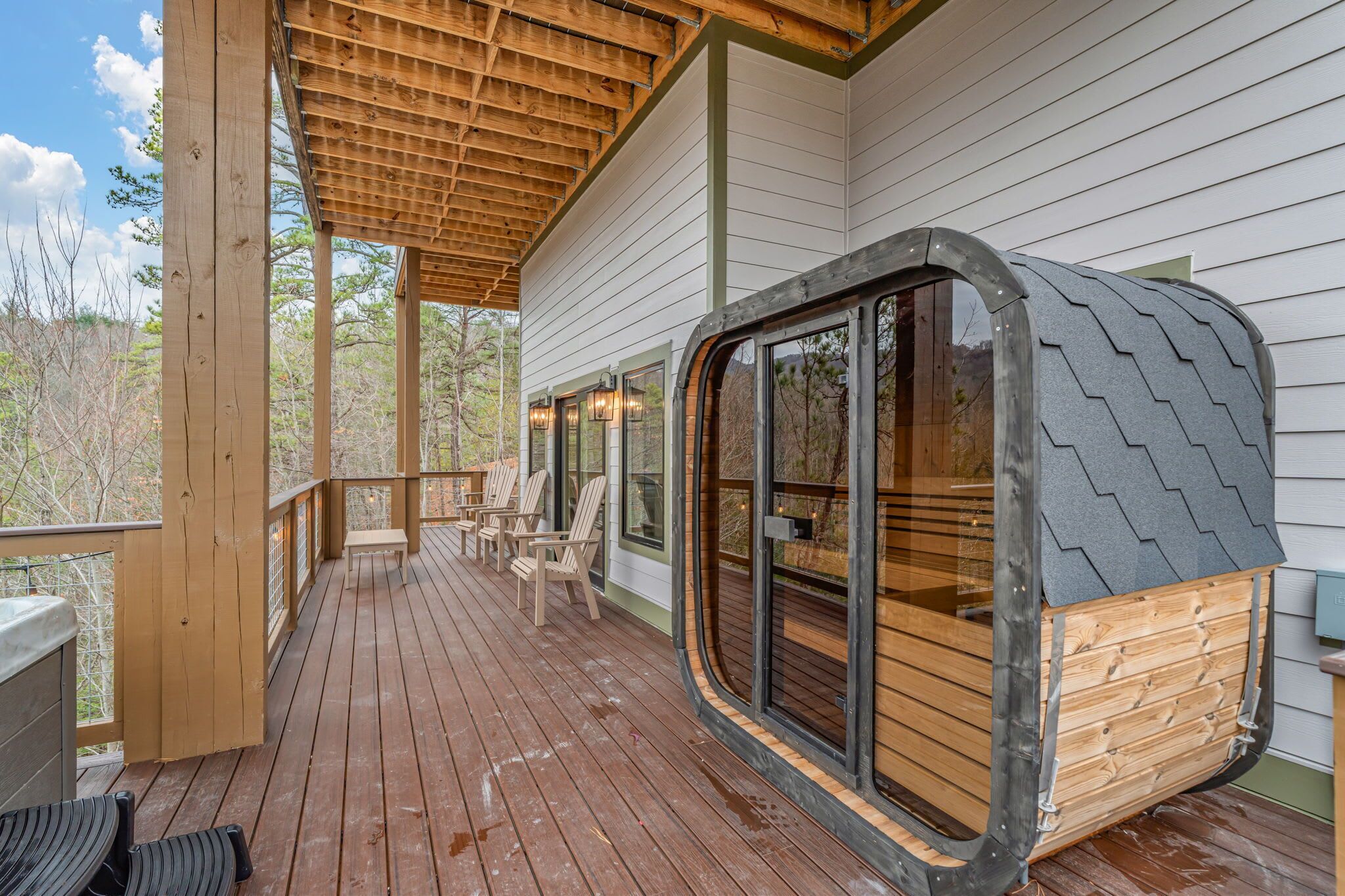 Una terraza de madera con un espacio de estar cubierto, una pequeña construcción tipo cabaña con ventanas amplias, y vistas a los árboles circundantes.
