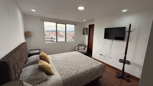 Ett sovrum med en säng, en tv som är monterad på väggen, ett fönster med utsikt över staden och ett nattduksbord med en lampa.