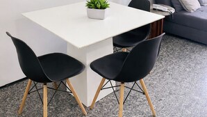 Ein modernes Wohnzimmer mit einem weißen Esstisch, zwei schwarzen Stühlen, einem grauen Sofa und einem kleinen Topfpflanzen.