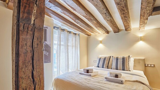 A bedroom with a wooden ceiling, a bed with striped pillows, and a window with curtains. - Amazing Apartment in Center of Paris (Paris)