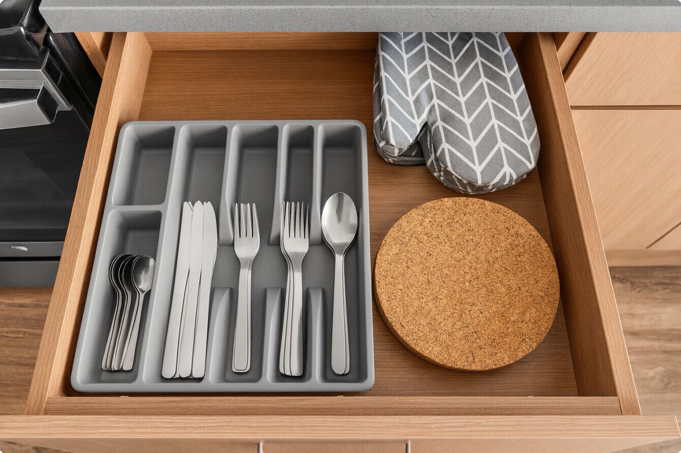 A drawer containing a gray silicone utensil tray with forks, knives, and spoons, a cork coaster, and a gray and white chevron-patterned oven mitt.