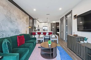 A modern living room with a green sofa, a dining table with red chairs, a kitchen area, and a TV mounted on the wall. - Modern Condo for 6 with Rooftop & Gym, Near DT! (Nashville)