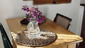 A wooden dining table with a woven placemat, a glass vase with purple flowers, and a window with wooden shutters. - Off grid Casa Rural MOZa LUZe (El Pilar)