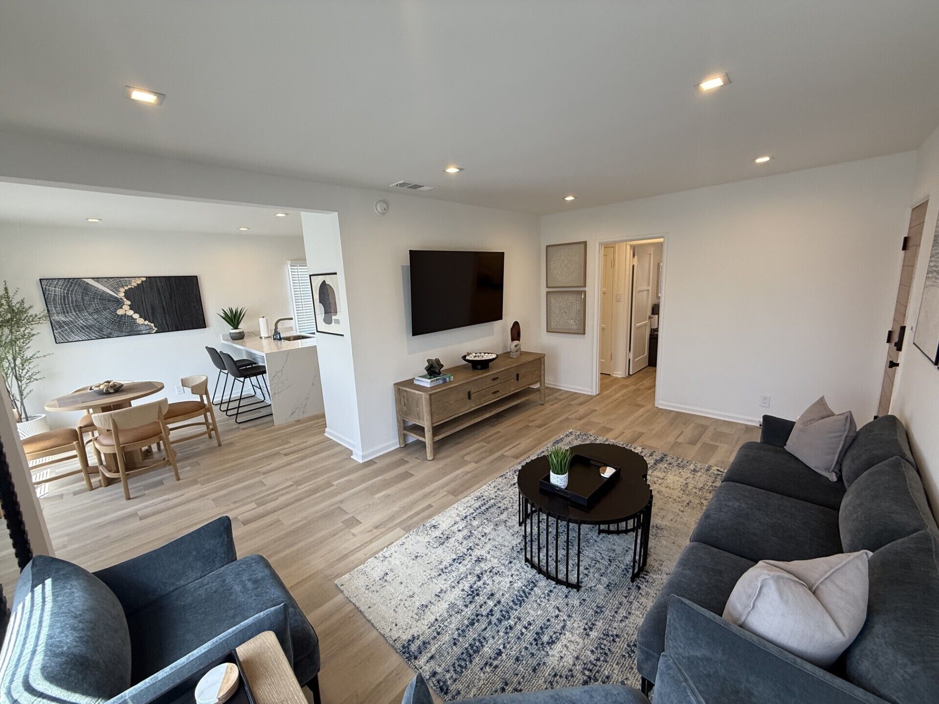 A modern living room with a sofa, coffee table, and TV. There is a dining area with a table and chairs. The room has wooden flooring and white walls.