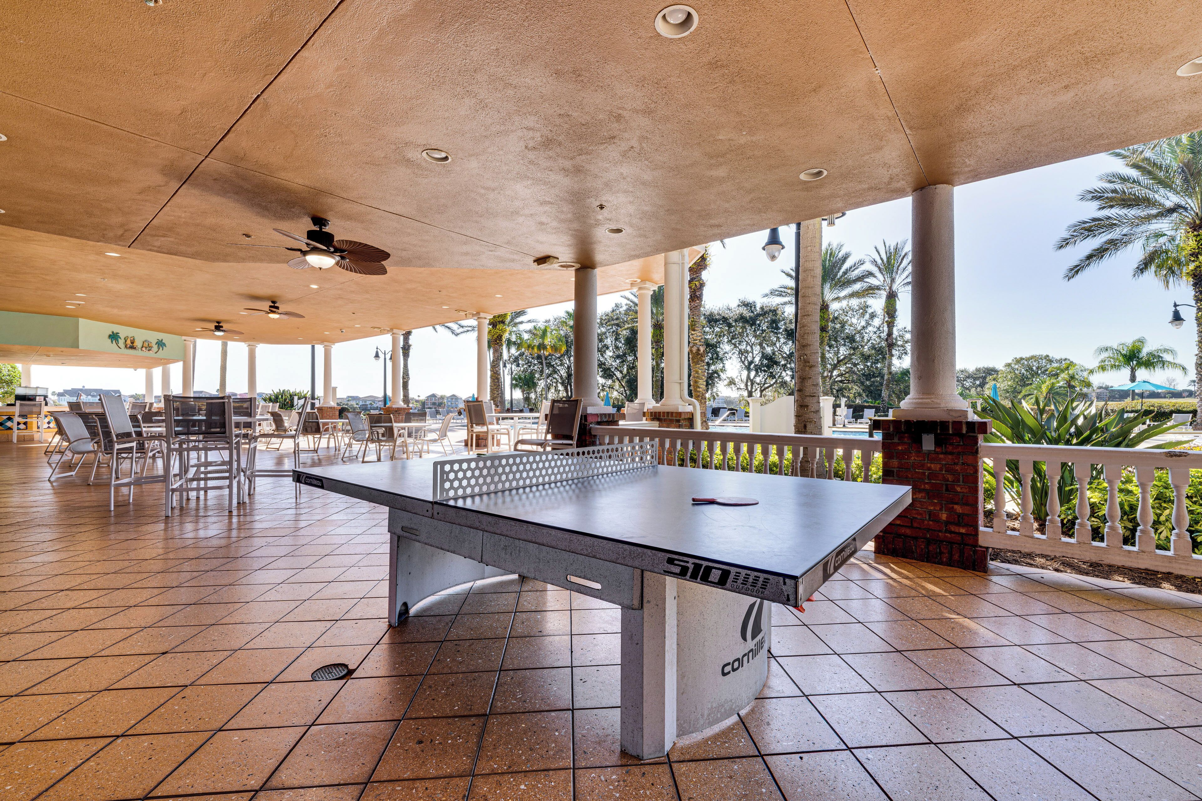 Una mesa de tenis de exterior con base de concreto, rodeada de sillas y un área de estar cubierta con vistas a palmeras y follaje.