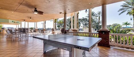 Una mesa de tenis de exterior con base de concreto, rodeada de sillas y un área de estar cubierta con vistas a palmeras y follaje.