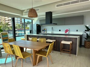 A modern kitchen with a central island, a dining area with a wooden table and yellow chairs, and a large window offering a view of the city. - Oceanfront Bucerias Escape | Huge Balcony & Panoramic Views 🌊 (Bucerías)