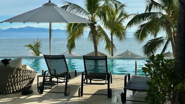 Un espace aménagé au bord de la piscine, avec des chaises longues et des parasols, des palmiers et une vue sur l’océan.