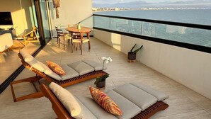 Un balcon doté de deux chaises longues, d’une table à manger et de chaises, offrant une vue sur une étendue d’eau et sur la terre ferme au loin.