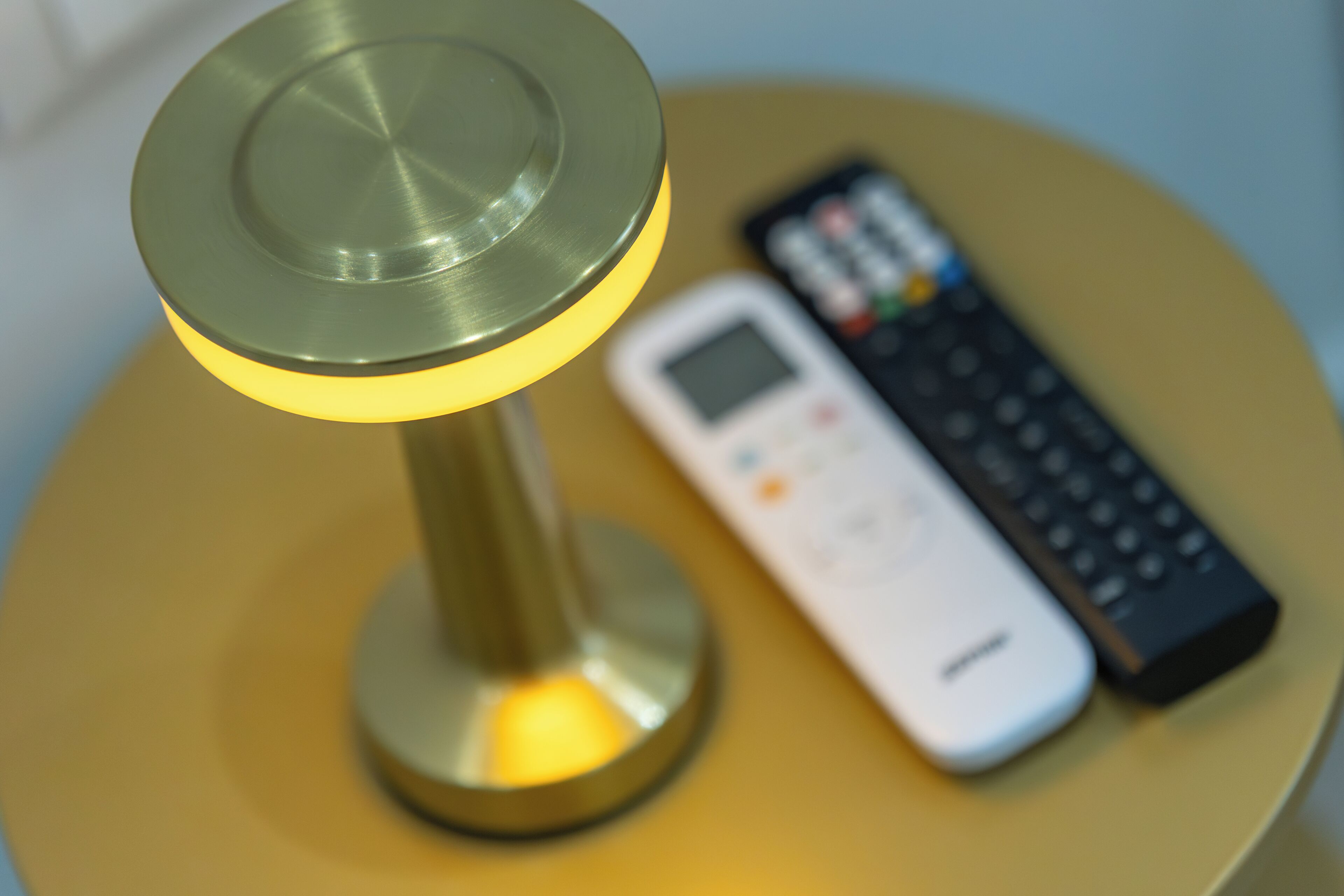 A lamp with a metallic base and a lit top, a white remote control, and a black remote control on a yellow surface.