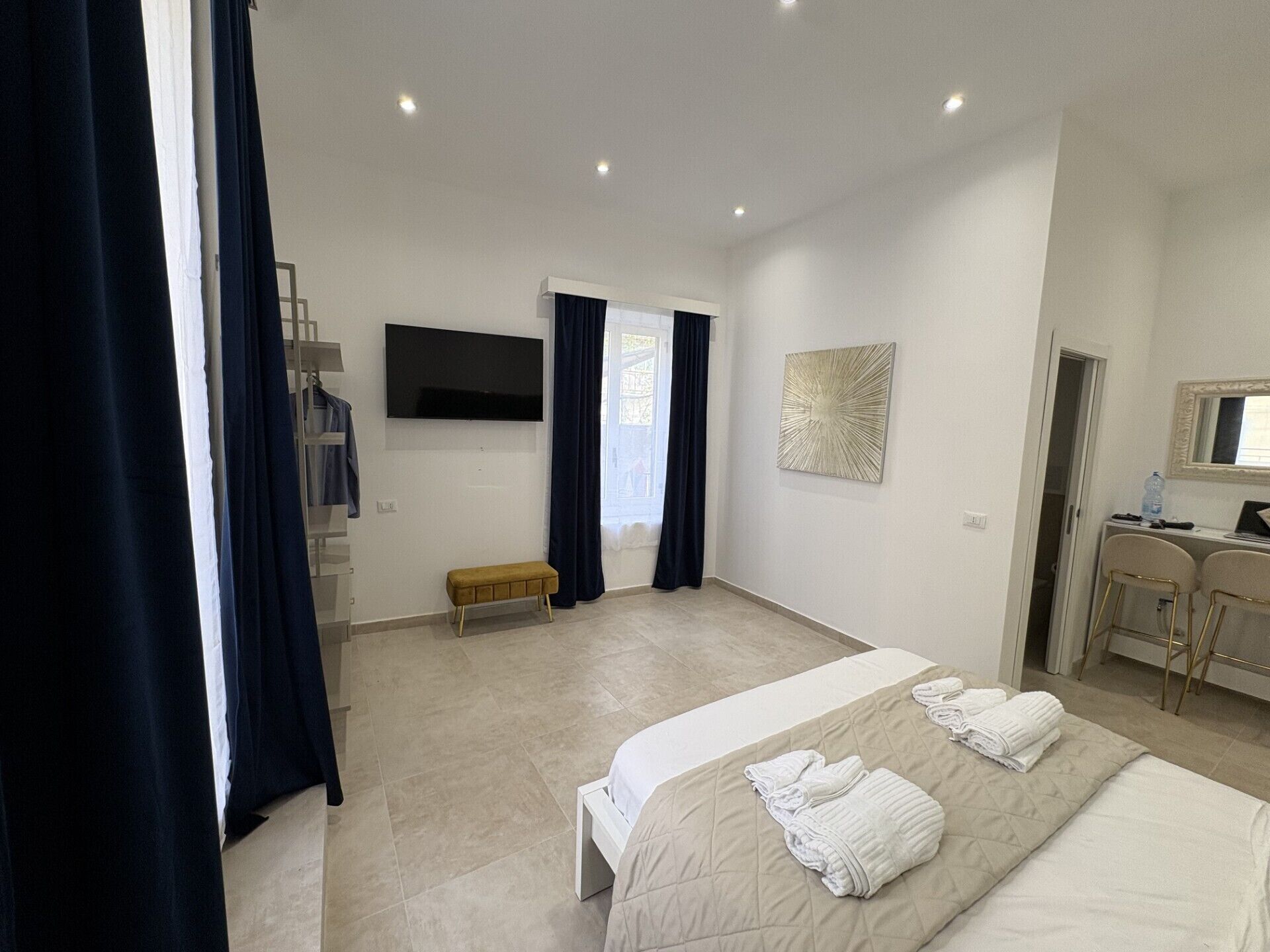 A hotel room with a bed, a bench, a TV, a desk with chairs, and a window with curtains.