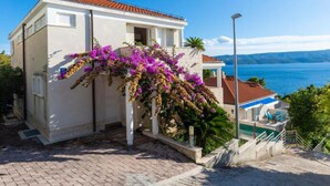 Ein weißes Gebäude mit rotem Ziegeldach, einem Balkon mit violetten Blumen und Blick aufs Meer.