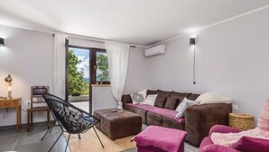 Ein modernes Wohnzimmer mit einem braunen Sofa, einem schwarzen Sessel, einem pinken Hocker und einem weißen Sessel. Ein Fenster mit Vorhängen, ein Schreibtisch mit Lampe und eine Wand-Klimaanlage sind ebenfalls zu sehen.
