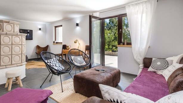 Ein modernes Wohnzimmer mit einer Couch, zwei Sesseln, einem kleinen Tisch und einem Schrank. Eine Glastür öffnet sich zu einem Balkon mit Blick auf Bäume.