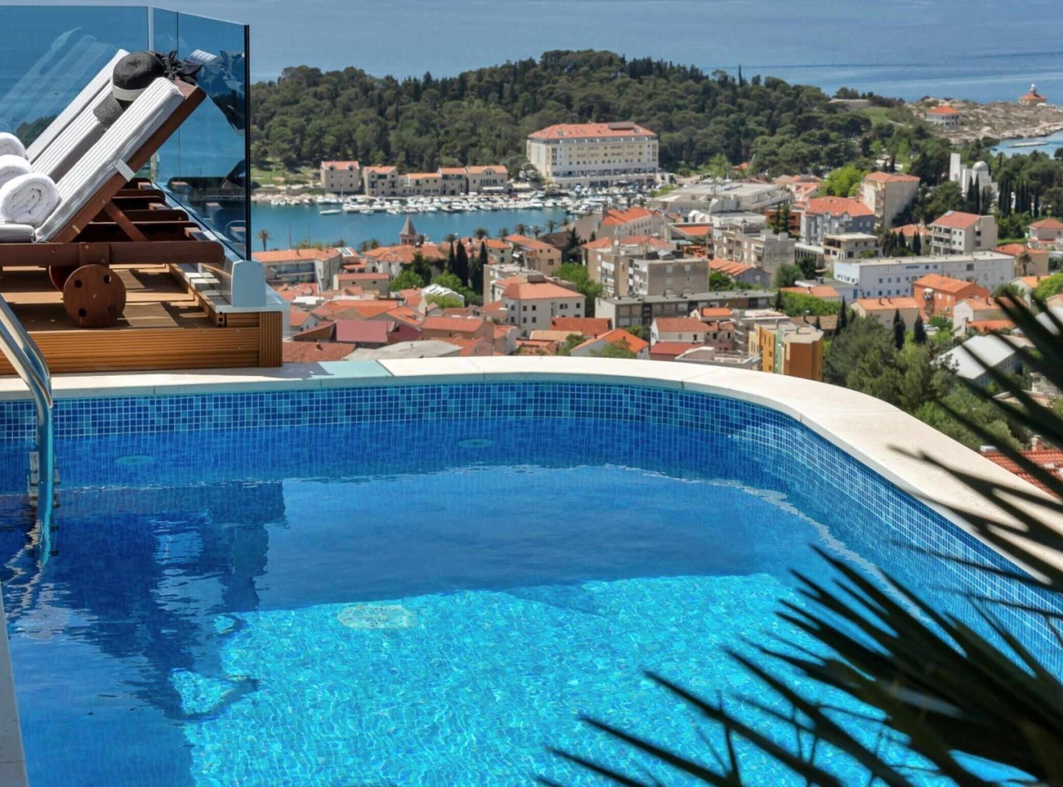 A swimming pool with clear blue water, lounge chairs, and a view of a coastal town with buildings and a hill in the background.