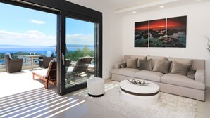 A modern living room with a large sliding glass door leading to a balcony, a beige sofa, a coffee table, and a painting of a sunset. - Lux Villa Afrodita Makarska - Three Bedoom Villa with Outdoor Pool (Makarska)
