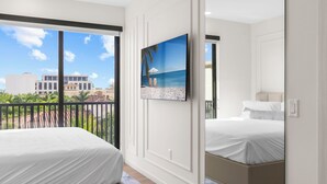 Une chambre dotée d’une grande fenêtre, d’un lit, d’un téléviseur fixé au mur et d’un balcon offrant une vue sur les immeubles et les palmiers.