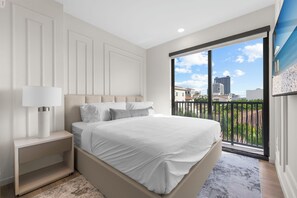 A bedroom with a bed, nightstand, and a view of buildings through a balcony.