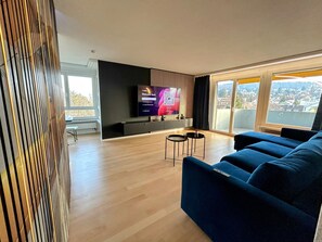 Un salon moderne avec un grand téléviseur à écran plat fixé au mur, un canapé bleu, une petite table basse et une vue sur le paysage urbain à travers de grandes fenêtres.